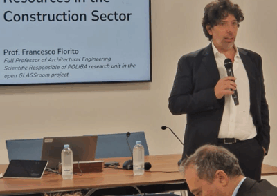 The picture shows Francesco Fiorito. He is holding a microphone and speaking to an audience. Behind him is a screen displaying the words “Open Educational Resources in the Construction Sector.”