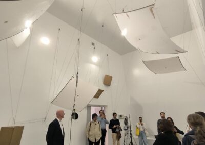 A small crowd of people stands inside a white room that has a very high ceiling. Metal rectangles are attached to the walls and ceiling with wires and hang above the people.