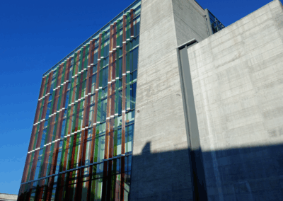 A building belonging to the Politecnico di Bari. The building is mostly made of concrete, but one façade is entirely made of glass. Green, orange, and red glass panels are attached to the facade, shining brightly in the sunlight.